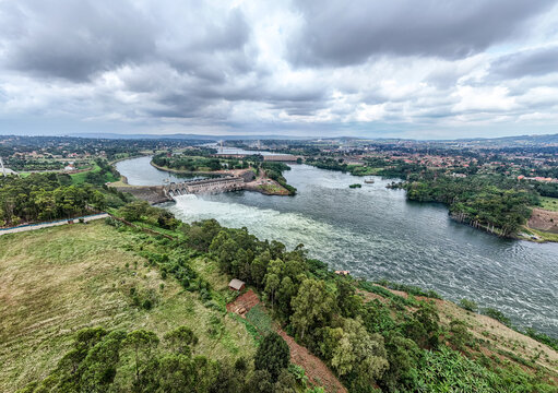 Aerial view of Owen Falls Dam with water flowing through the spillway into the river, surrounded by lush green landscape and a cloudy sky in Jinja, Eastern Region, Uganda.