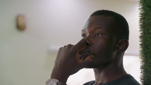 Black man waiting in intimate interior, chin resting on hand and watch visible soft light, subtle grainy film look, greenery blurred in background, thoughtful gaze, rehearsing conversation, imagining