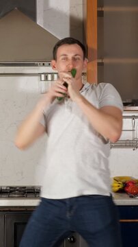 Positive man in a good mood having fun in his kitchen, actively dancing and singing into a cucumber instead of a microphone, expressing happiness, freedom, and enjoyment of life