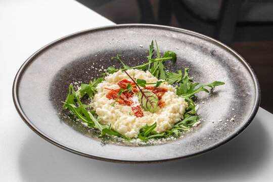 Creamy risotto served on a black plate garnished with arugula, sun-dried tomatoes, and herbs on a white table in a modern dining setting