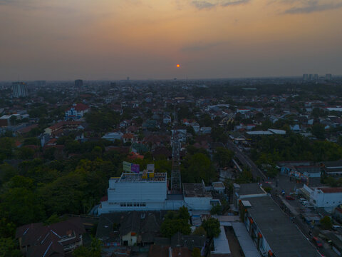 Aerial view of Condong Catur at sunset, featuring dense urban rooftops, a communication tower, and a hazy orange sky over the city landscape in Depok, Special Region of Yogyakarta, Indonesia.