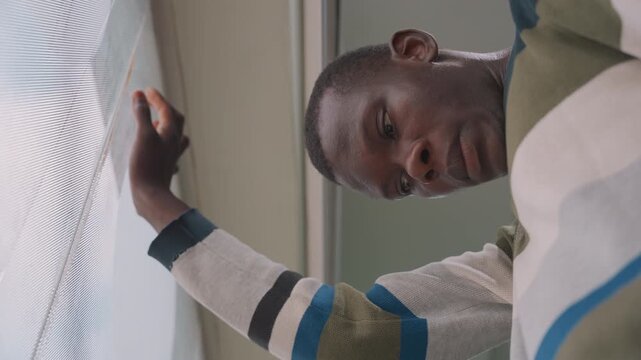 Youthful student gazes. Teenager in striped clothing rests hand on blinds enjoying peaceful moment. Young black individual with gentle eyes leans by window during quiet afternoon in cozy apartment