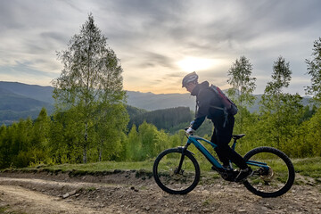 Fototapeta premium Side view of mountain biker riding electric bike on dirt trail through green forest hills at sunset, scenic landscape and outdoor adventure concept