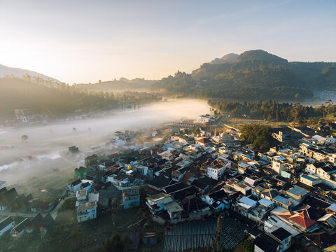 Aerial view of Dieng village nestled in a valley filled with morning mist, surrounded by green mountains and terraced fields under golden sunlight in Kejajar, Central Java, Indonesia.