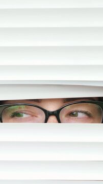 Suspicious woman wearing glasses peeking through opening horizontal blinds, her eyes furtively looking around, conveying a sense of curiosity, spying, and paranoia in a close up shot