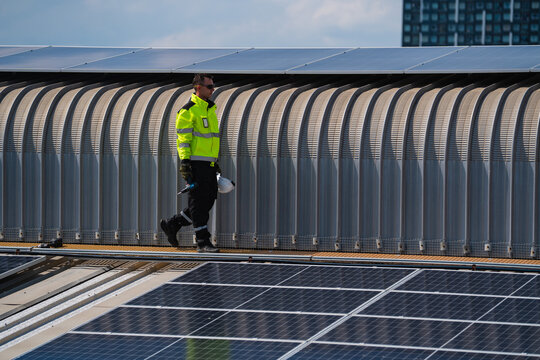 Solar panel worker rooftop safety helmet industrial energy maintenance high visibility urban Worker walking rooftop near solar panel array wearing high visibility jacket and helmet for industrial