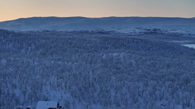 Drohne fliegt &uuml;ber einer Schneelandschaft in einem finnischen Dorf hinter dem Polarkreis w&auml;hrend einer Polarnacht. Wald mit schneebedeckten B&auml;umen. Kleine H&auml;user im Schnee. Gem&uuml;tlichkeit und Lichter.