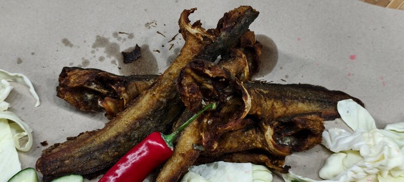 Deep-fried catfish (ikan keli goreng in Malay or lele goreng in Indonesian), golden-brown and crispy, served with fresh raw cabbage and a red chili pepper on brown paper.