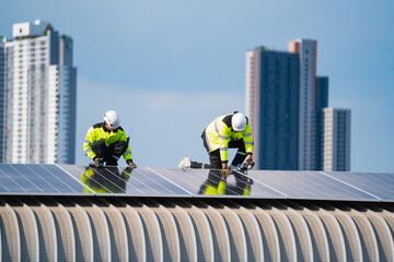 Fototapeta premium Solar panel worker rooftop helmet safety vest city building renewable energy installation, two construction workers installing photovoltaic panels urban roof with high rise skyline clear blue sky