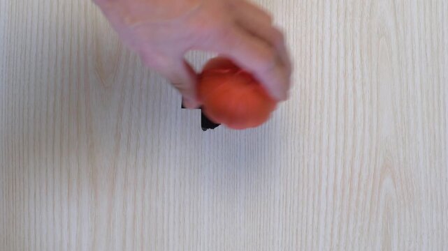 Overhead hand rolling orange ball across tape marker on light wood surface, calibration and alignment test, checking path and balance, gentle motion for placement accuracy