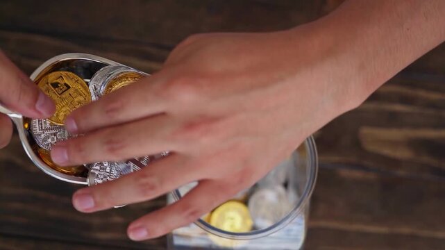 Hands carefully transfer gold and silver bitcoin tokens into a transparent glass jar placed on a dark wooden table as a metaphor for long term financial planning and systematic wealth building.