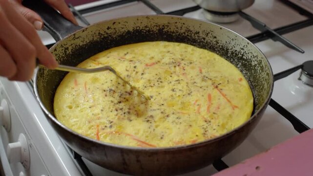 Close-up kitchen scene showing hands using fork to lift, fold, and shape cooked omelet inside seasoned cast iron skillet on gas stove, with warm rustic texture and appetizing breakfast focus