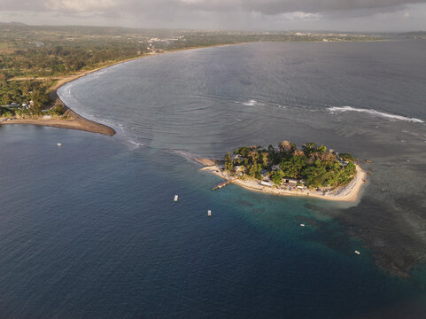 Aerial view of a small tropical island with lush vegetation, a wooden pier, and small boats anchored in the turquoise waters near the coastline of Port Vila, Shefa Province, Vanuatu.