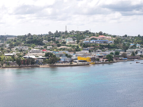 Aerial view of the coastal cityscape of Port Vila featuring buildings, lush green hills, and clear blue water in the harbor under a cloudy sky in Port Vila, Shefa Province, Vanuatu.