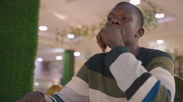African man enjoying meal. Individual sampling food at cafe. Man with striped clothing tastes dish at mall restaurant. Gentleman with hand over mouth savors meal during casual date in mall