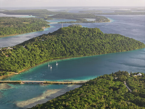 Aerial view of Vava'u islands featuring lush green hills, turquoise waters, a bridge connecting landmasses, and sailboats anchored in the bay in Vava'u, Tonga.