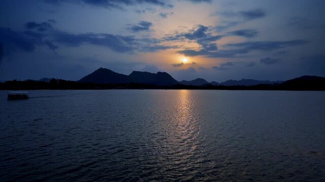 Golden light streams across rippling water as dusk falls over distant peaks. A small boat moves along the quiet horizon. Evening skies glow above Udaipur�s tranquil lake.