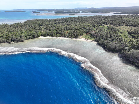 Aerial view of the tropical coastline of Vava'u, featuring a vibrant coral reef, turquoise shallow waters, white sandy beach, and dense palm tree forest in Vava'u, Tonga.