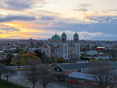 Aerial view of Sacred Heart Basilica surrounded by urban rooftops and trees under a dramatic sunset sky in Timaru, Canterbury Region, New Zealand.