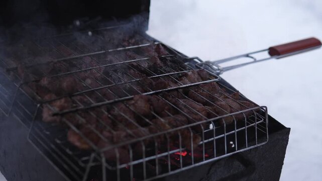 A close-up, slightly elevated view of cooking meat on a grill in a cozy outdoor winter setting with warm tones and metal and wood materials. The scene features brown and red colors, and itp.