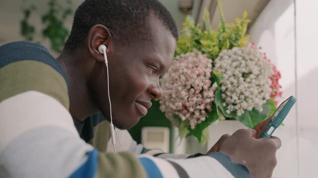 Black man on video date smiling, wearing earbuds, listening intently, laughing softly and typing affectionate replies while sitting by window with bouquet of hydrangea, warm daylight and cozy sweater