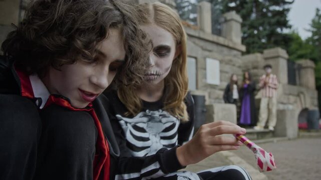 Caucasian teenagers wearing Halloween costumes sitting outdoors, sorting candies from orange bucket. Clip is ideal for seasonal holiday marketing and Halloween promotions
