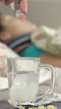 Close up of a hand pouring medicinal powder from a sachet into a glass of water and stirring it, preparing a remedy for a sick person resting in the background on a bed