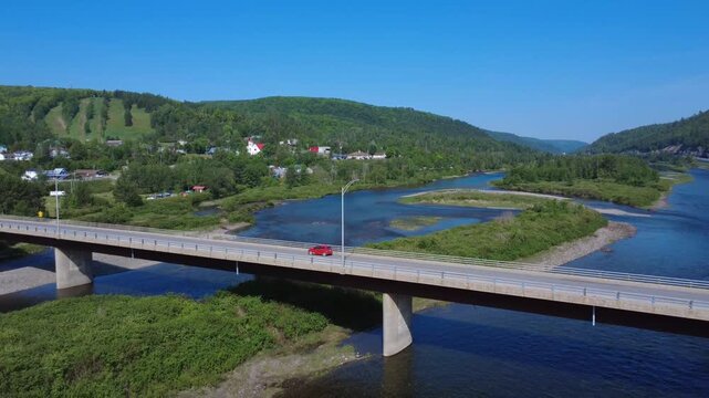 Drone footage captures a red car crossing a bridge over the scenic Matapedia River surrounded by forests and mountains in Quebec, Canada.