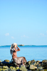 Full-length side profile portrait of a young Southeast Asian Muslim woman wearing a beige hijab and brown long-sleeved dress, sitting gracefully on rocks at the beach. Features calm blue ocean waters, © Slaf