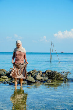 ​A young Asian Muslim woman in a hijab holding her skirt while standing in clear, shallow ocean water near a rocky shore under a bright blue sky. Concept of tropical vacation, modesty travel, peaceful