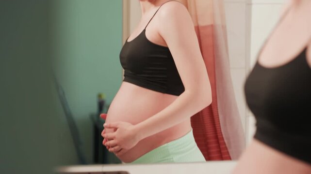 Pregnant woman examining bump in mirror, soft morning light and black tank top, hands gently cradling belly, checking skin and posture, practicing breathing and prenatal selfcheck, quiet domestic
