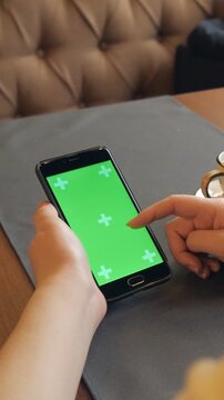 Woman's hand tapping and scrolling on a smartphone with a green screen display in a cafe, over the shoulder perspective showing digital interaction and mobile technology concept