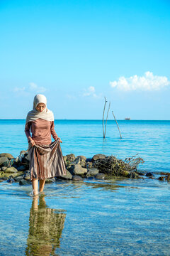 ​A young Asian Muslim woman in a hijab holding her skirt while standing in clear, shallow ocean water near a rocky shore under a bright blue sky. Concept of tropical vacation, modesty travel, peaceful