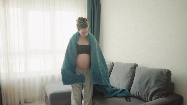 Pregnant white woman wrapping blanket over bare belly in sunlit living room, nesting on gray sofa with teal throw, slow gentle movements, pajama pants, relaxed maternal expression, intimate selfcare