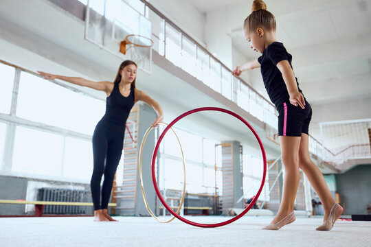 Young girl jumping beside hoop while female gymnastics coach guides exercise. Concept of rhythmic hoop training, jumping coordination, confidence in beginner gymnastics.