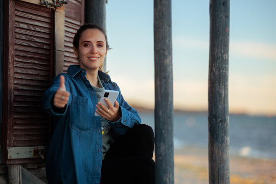 Happy Woman In Denim Jacket Making Thumbs Up At Sea. Cheerful tourist holding smartphone and showing positive gesture 