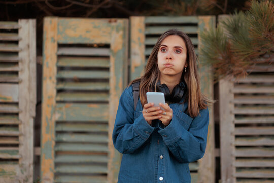 Puzzled Woman In Denim Jacket Holding Phone Outside Old Restaurant. Doubtful female tourist wearing sweater and headphones looks at smartphone outside deserted building