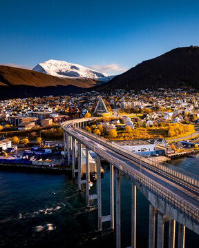 Aerial view of the Arctic Cathedral and the bridge leading into the town of Tromsdalen with a snow-capped mountain in the background under a clear blue sky in Tromsdalen, Troms, Norway.