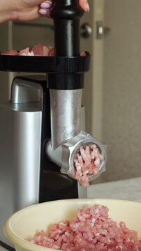 Female hands pushing fresh raw meat cubes into a modern electric meat grinder, processing pork and beef into fresh minced meat falling into a bowl for a homemade meal
