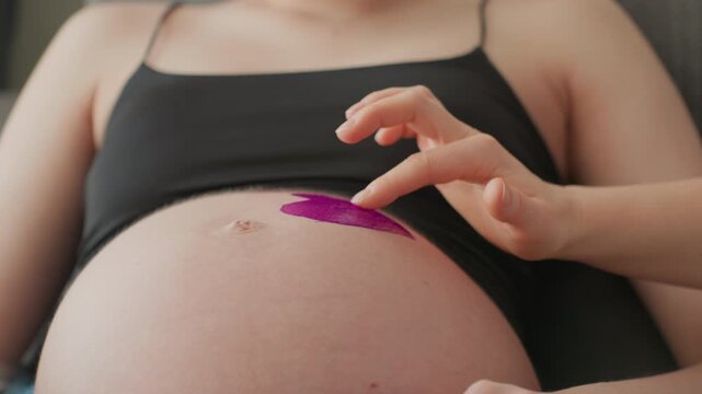 Pregnant woman painting purple heart on belly, closeup of exposed bump with black crop top, gentle fingertip strokes applying vivid purple paint, soft natural light, quiet cozy bedroom atmosphere,