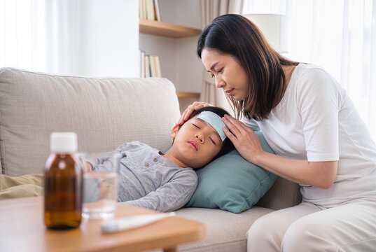 Worried Asian mother checking the temperature of her sick boy sleeping on the couch with a cooling patch on his forehead