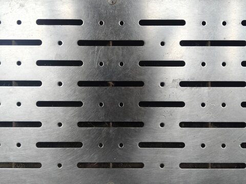 Industrial metal drainage cover featuring repetitive geometric slot patterns and circular holes designed for heavy duty urban infrastructure flooring and effective water runoff systems.