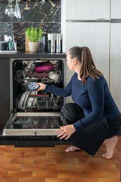 A young woman loads plates into the rack of an open dishwasher in a modern kitchen with marble and dark cabinets a candid domestic scene of everyday household chores.