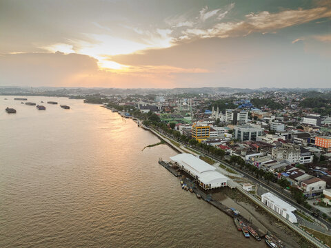 Aerial view of the Mahakam River flowing past Samarinda at sunset, with cargo barges on the water and urban buildings along the riverbank in Samarinda, Kalimantan Timur, Indonesia.
