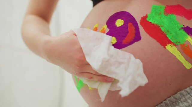Pregnant belly covered with colorful paint, playful handprint and abstract strokes across skin, closeup of expectant mother using tissue to wipe paint, warm bathroom light, joyful creative maternity