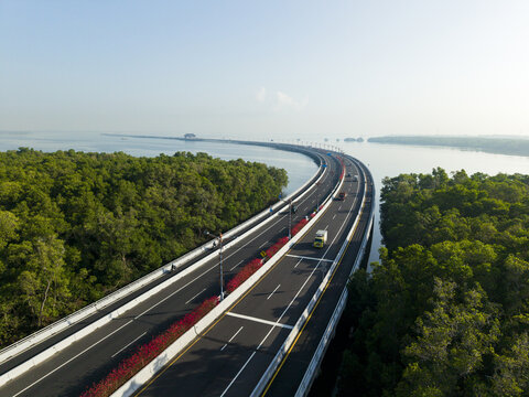 Aerial view of Mandara Toll Road stretching over the sea, surrounded by lush mangrove forests, with vehicles driving on the highway in Kecamatan Kuta Selatan, Bali, Indonesia.