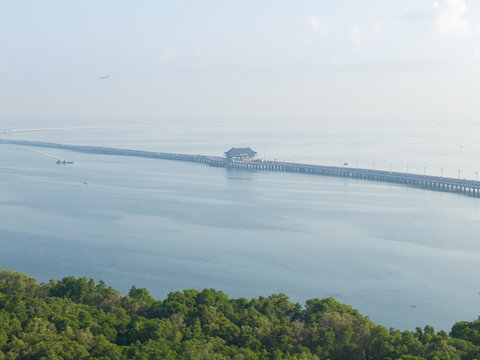 Aerial view of Mandara Toll Road stretching across the calm bay waters, surrounded by lush green mangroves under a clear sky in Kecamatan Kuta Selatan, Bali, Indonesia.