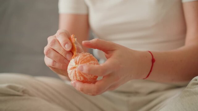 Closeup hands peeling orange on lap, delicate fingers separating segments, juice droplets, red string bracelet, calm domestic setting, slow mindful motion, tactile texture, wellness snack, sensory