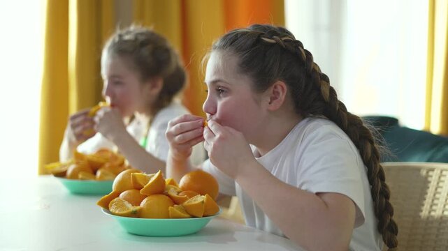 Girl eating orange slices at table enjoying healthy citrus fruit. Child with braid eats fresh orange segments. Kids share vitamin snack indoors. Children enjoy nutritious fruit together.
