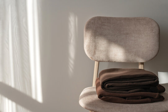 Brown jersey clothes on beige chair near empty beige wall background with abstract natural sunlight shadows, minimal cozy home interior still life, slow living